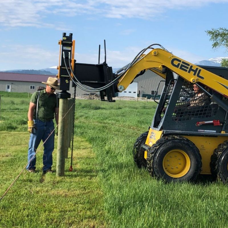 skid steer driving wooden post in the field with a montana post driver
