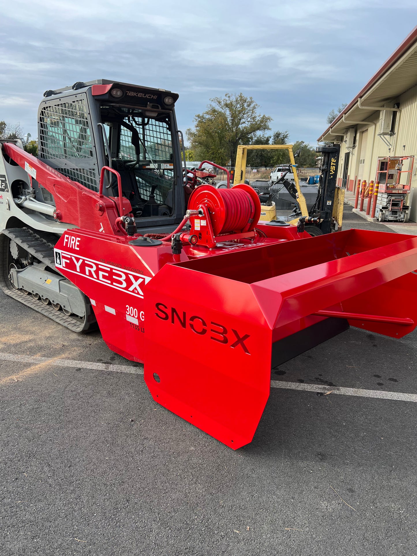 Red FYREBX SNOBX snow and debris attachment mounted on a track loader parked outside a shop