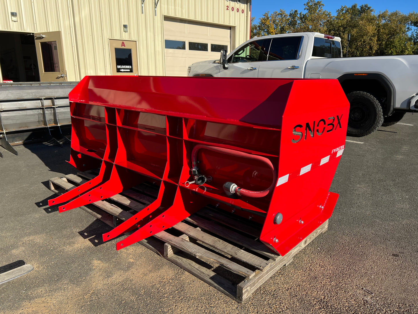 Red SNOBX FYREBX attachment with hydraulic ports resting on a pallet in a parking lot near a shop and truck