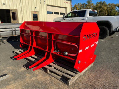 Red SNOBX FYREBX attachment with hydraulic ports resting on a pallet in a parking lot near a shop and truck