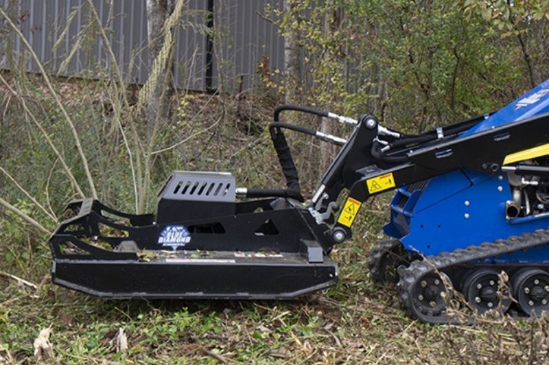 Mini skid steer with the open front brush cutter attachment cutting small trees in the forest by Blue Diamond