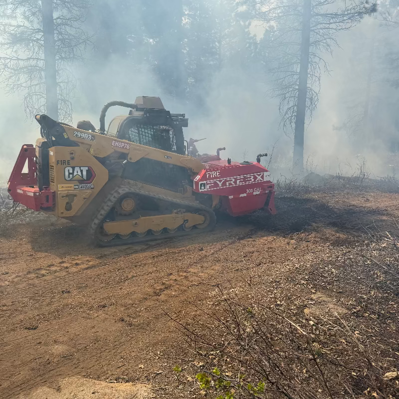 CAT skid steer with a FYREBX attachment working through smoke while controlling a small ground fire in a forested area