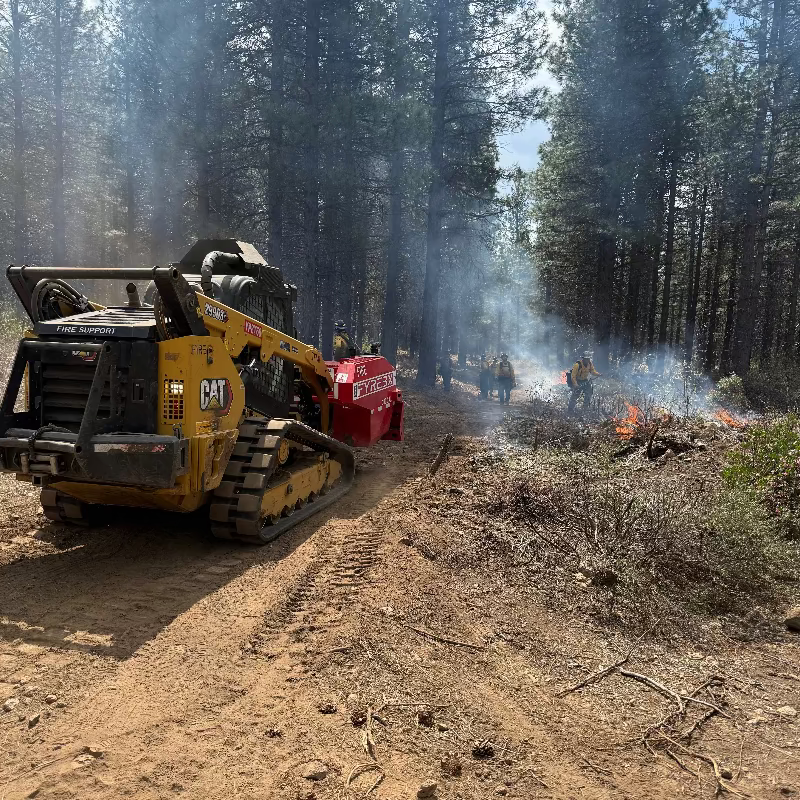 CAT skid steer with FYREBX attachment working near firefighters as smoke rises from a small ground fire in a forest