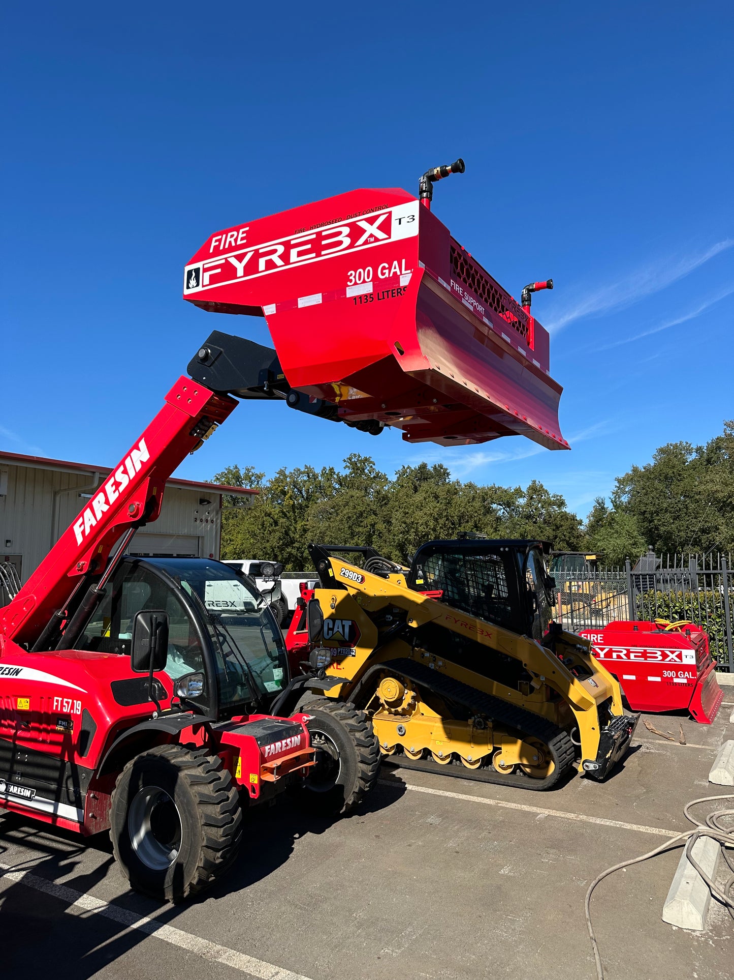 Red telehandler lifting a FYREBX 300-gallon firefighting attachment beside a CAT track loader in a sunny outdoor lot