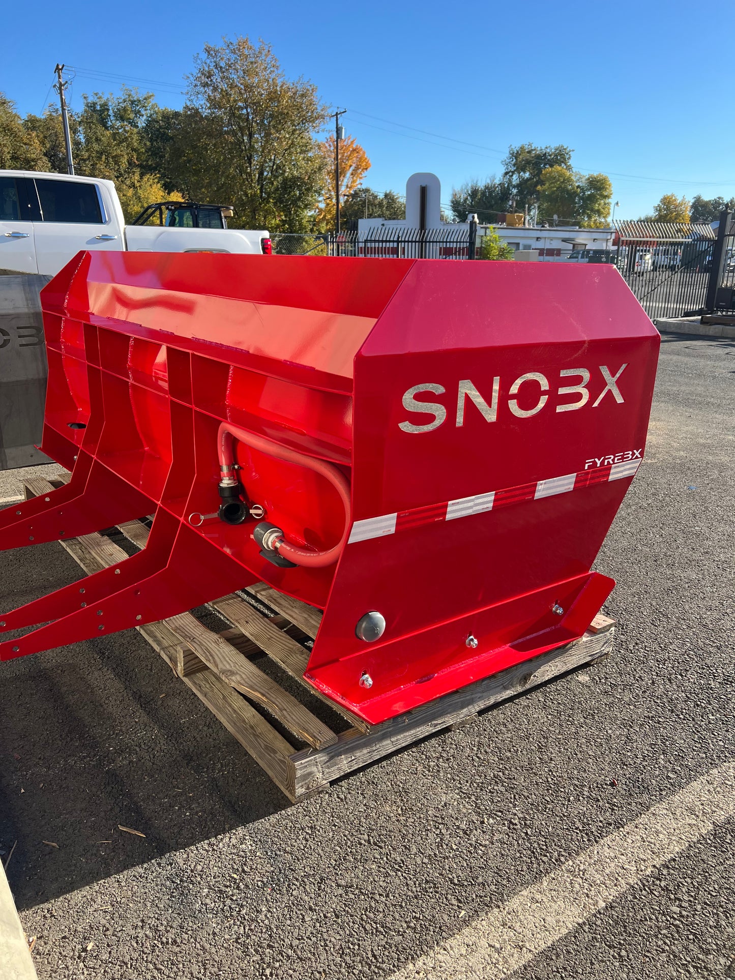 Red SNOBX FYREBX attachment with hydraulic lines, displayed on a pallet in an outdoor parking area